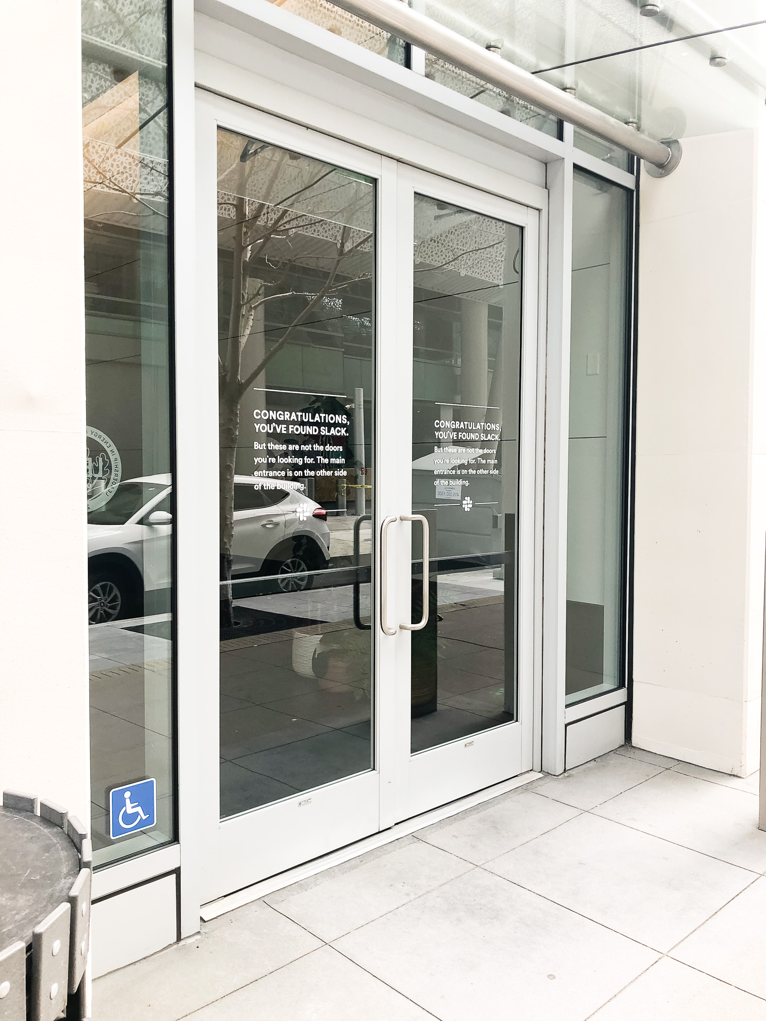 Matte white vinyl wayfinding sign with white text on glass doors for the San Francisco office of Slack, an American cloud-based set of proprietary team collaboration tools and services.