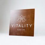 Dark wood retail blade sign with white artwork for Vitality, a massage therapy and wellness studio located in Carytown, Virginia.