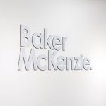 Brushed metal lobby sign on white wall at the office of Baker McKenzie, a multinational law firm.