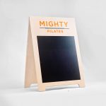 Light wood A-frame sign with orange logo and chalkboard area for class menu for Mighty Pilates, a fitness chain.
