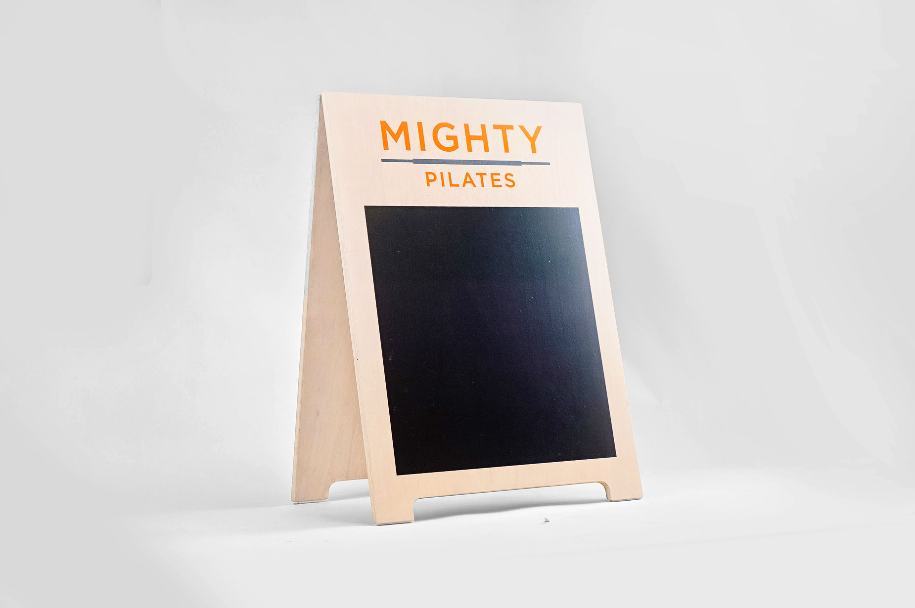 Light wood A-frame sign with orange logo and chalkboard area for class menu for Mighty Pilates, a fitness chain. Light wood A-frame sign with orange logo and chalkboard area for class menu for Mighty Pilates, a fitness chain.
