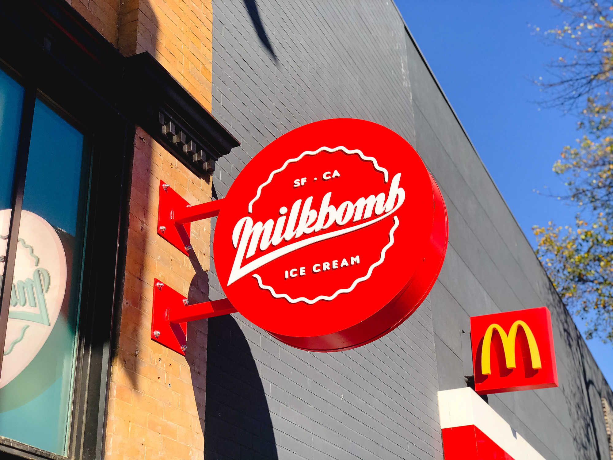 Red circular blade sign with white script for Milkbomb, a sweet stop for donut ice cream sandwiches. Red circular blade sign with white script for Milkbomb, a sweet stop for donut ice cream sandwiches.