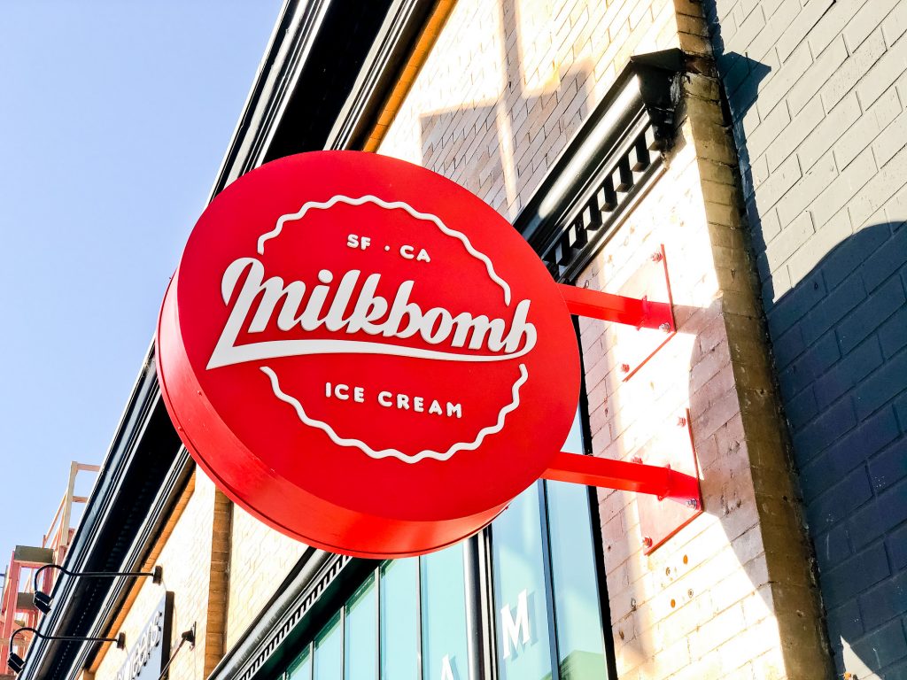 Red circular blade sign with white script for Milkbomb, a sweet stop for donut ice cream sandwiches.