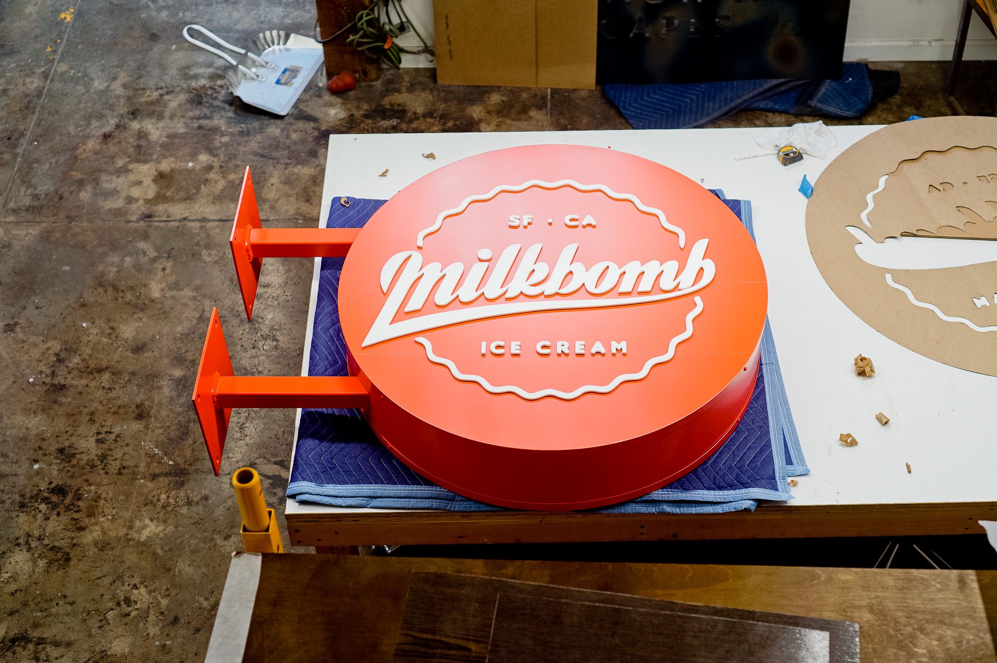 Red circular blade sign with white script for Milkbomb, a sweet stop for donut ice cream sandwiches.