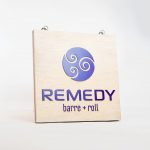 Hanging wood blade sign with purple logo for Remedy Barre and Foam Rolling, an independent studio offering barre & foam-roller stretching classes in Oakland CA.