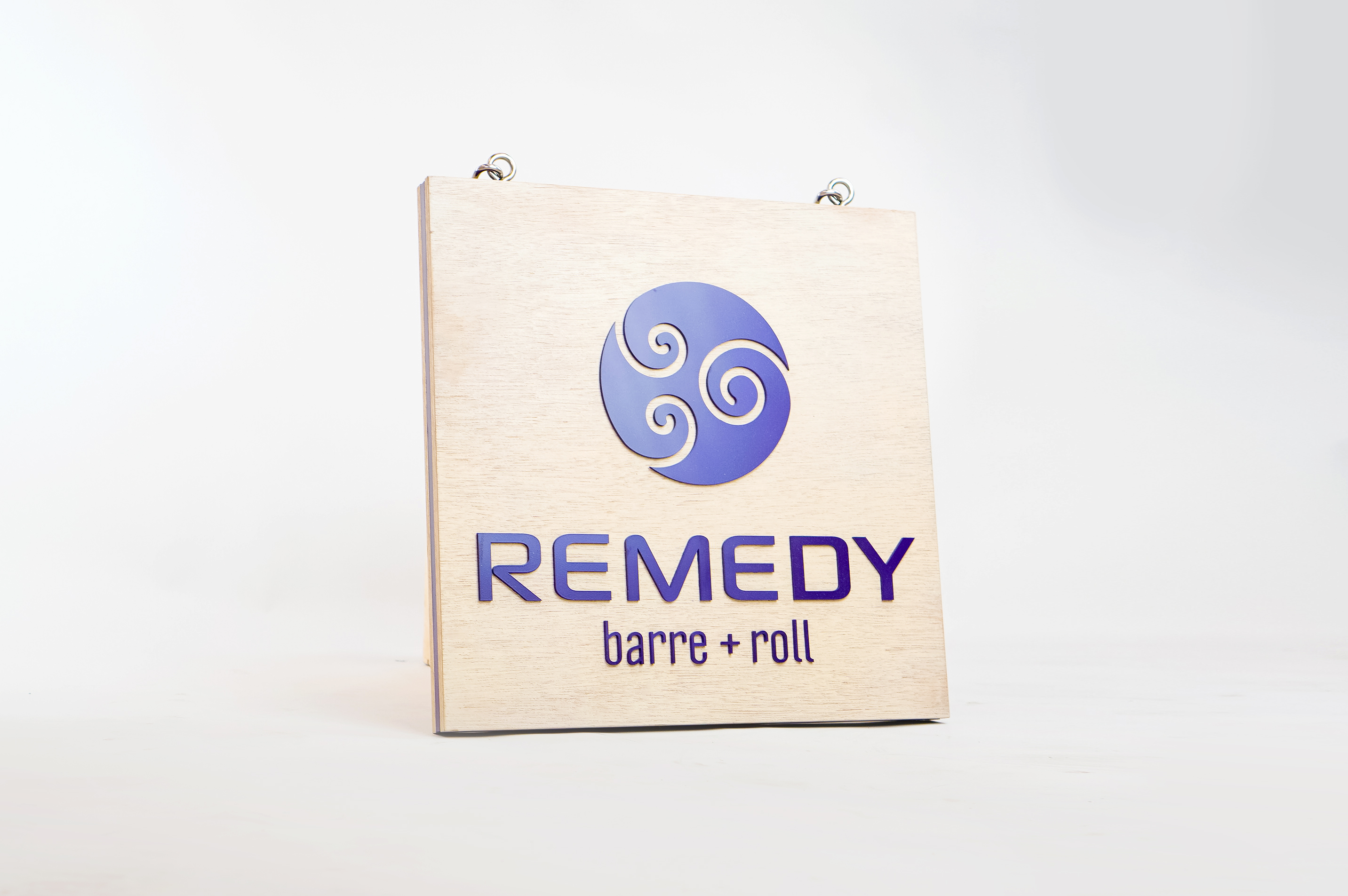 Hanging wood blade sign with purple logo for Remedy Barre and Foam Rolling, an independent studio offering barre & foam-roller stretching classes in Oakland CA.