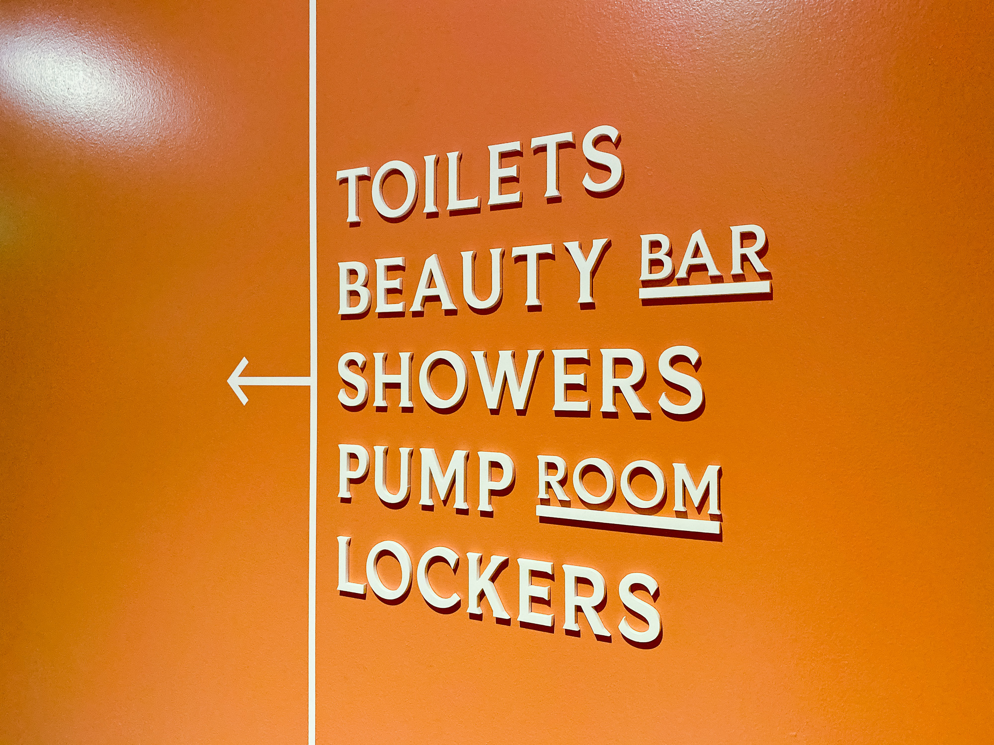 Cream colored, typographic wayfinding sign on orange wall for The Wing, a co-working space in San Francisco. Cream colored, typographic wayfinding sign on orange wall for The Wing, a co-working space in San Francisco.
