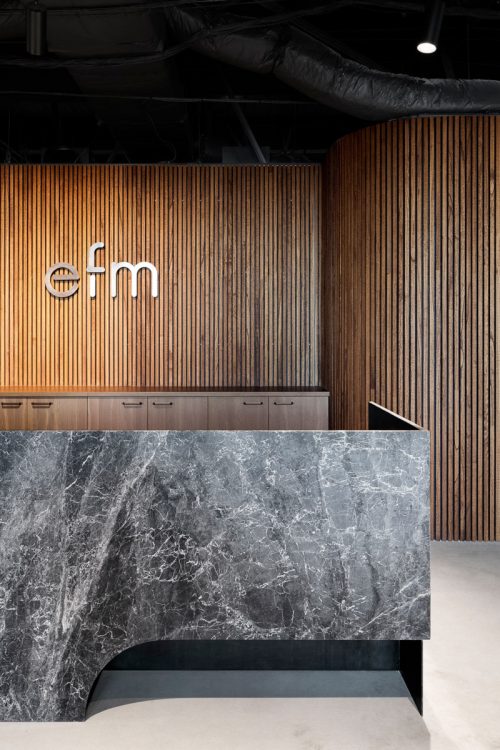 Metal logo on slat wood wall behind marble reception desk at the Melbourne office of EFM, a supply chain company. (Photo: SpaceCraft)