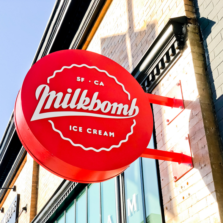 Red circular blade sign with white script for Milkbomb, a sweet stop for donut ice cream sandwiches.