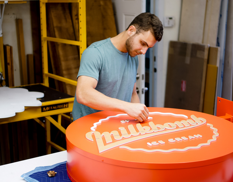 Red circular blade sign with white script for Milkbomb, a sweet stop for donut ice cream sandwiches.