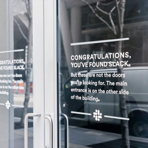 Matte white vinyl wayfinding sign with white text on glass doors for the San Francisco office of Slack, an American cloud-based set of proprietary team collaboration tools and services.